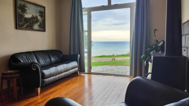 A living room with a black leather sofa, a wooden floor, a painting on the wall, and a view of the sea through a glass door.