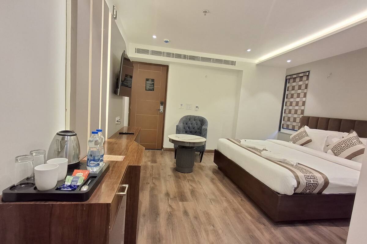 A hotel room with a wooden floor, a bed with patterned pillows, a desk with a kettle and cups, and a chair.