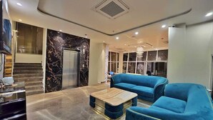 Un vestíbulo moderno con suelo de mármol, un conjunto de sofás azules, una mesa de centro de mármol y una escalera que lleva a un ascensor.