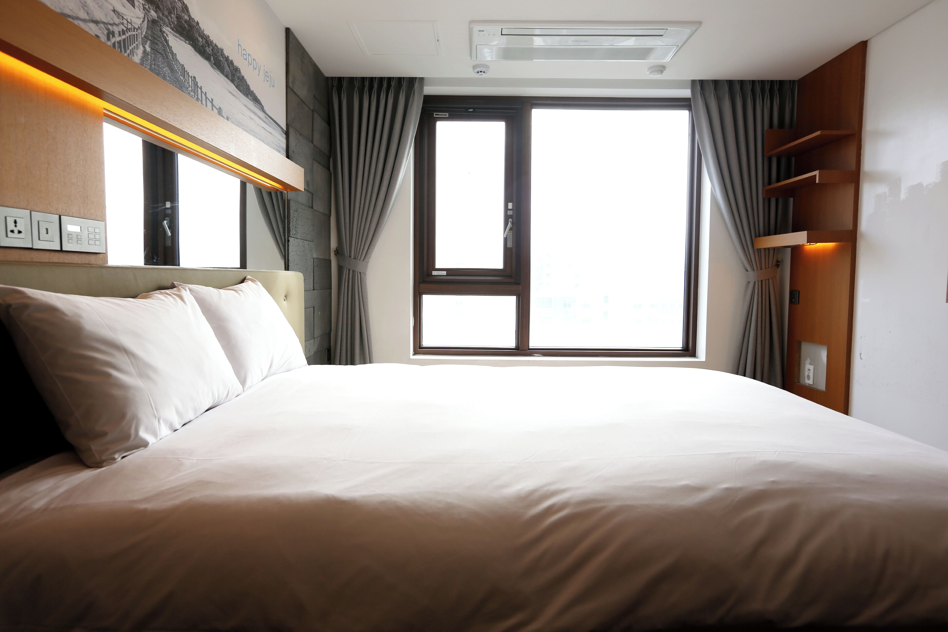 Hotelkamer met een groot bed, een raam met gordijnen, een wandairconditioner en een plank met een fles.