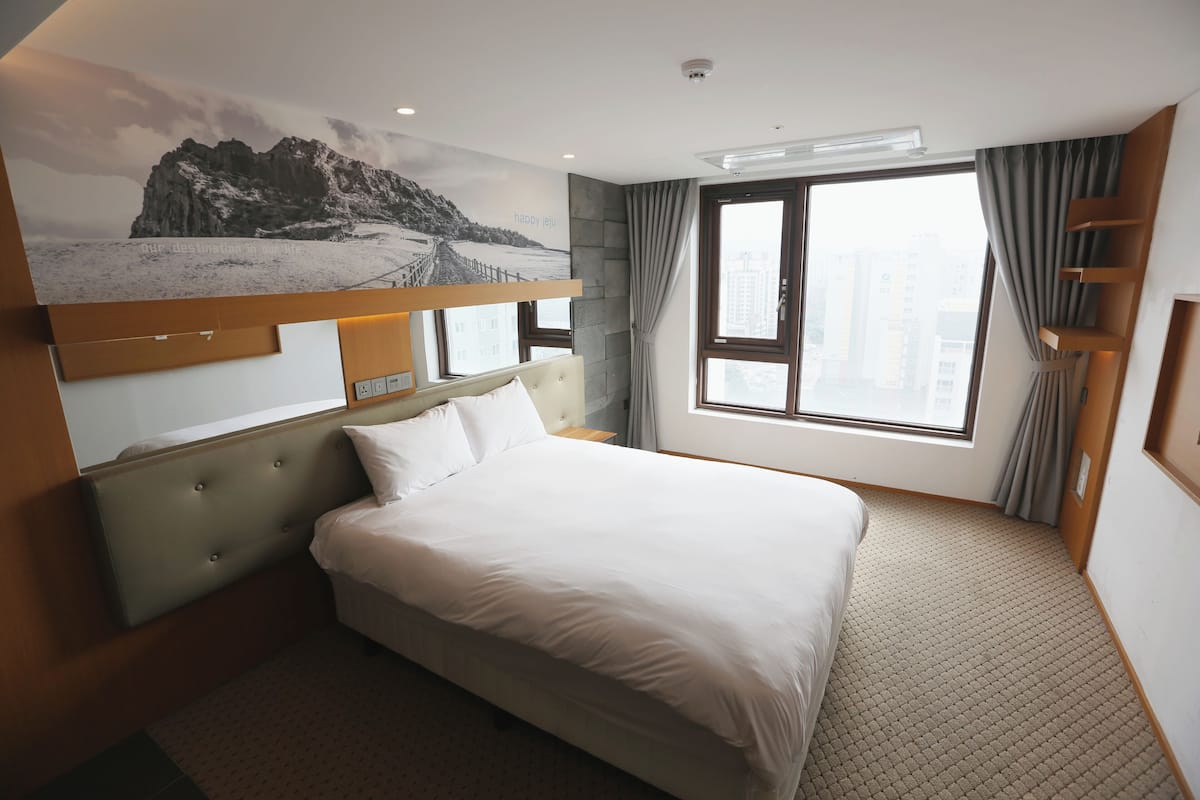 Een hotelkamer met een groot bed, hoofdbord, een raam met gordijnen, een wandplank en uitzicht op de stad.