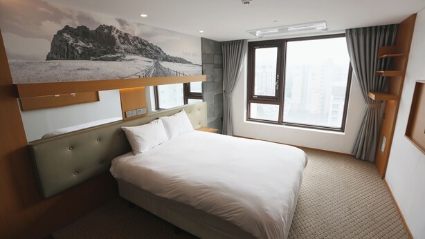 Een hotelkamer met een groot bed, hoofdbord, een raam met gordijnen, een wandplank en uitzicht op de stad.