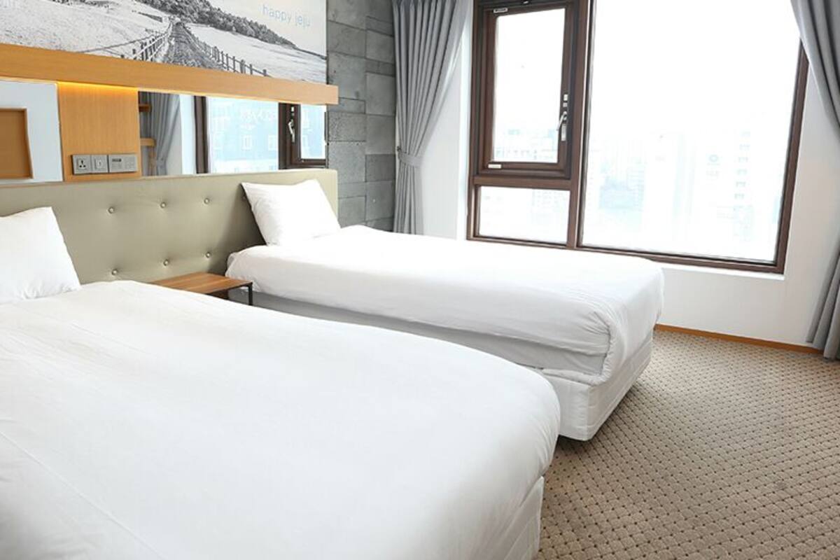 Een hotelkamer met twee bedden, een hoofdbord, een raam met gordijnen en uitzicht op de stad.