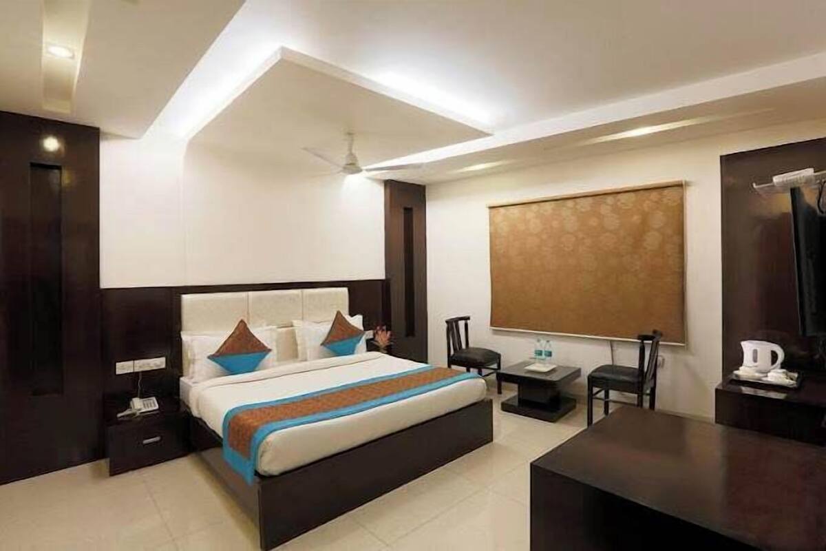A modern hotel room with a large bed, bedside tables, a desk, and a window with blinds.