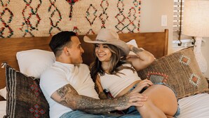 A couple lying on a bed with a patterned wall behind them, a wooden headboard, and a lamp on a bedside table. - Sacred Sands - Lobo Luna (Joshua Tree)