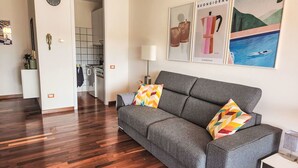 A modern living room with a grey sofa, colorful pillows, a small table with a lamp, and artwork on the wall. - Nice apartment for 3 guests with WIFI, A/C, TV, patio and pets allowed (Lesa)
