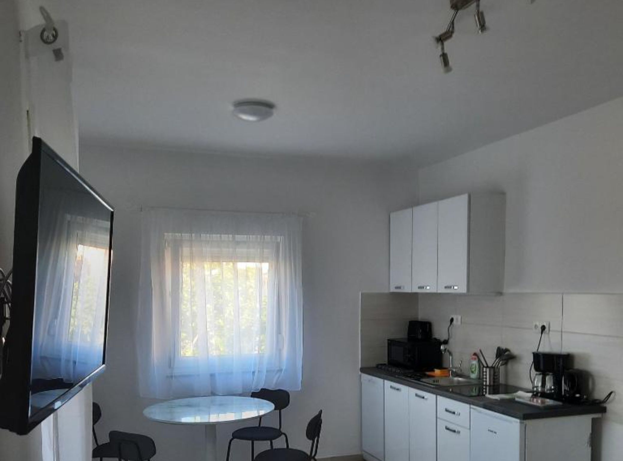 A compact kitchen with white cabinets, a small dining table, and a TV mounted on the wall.