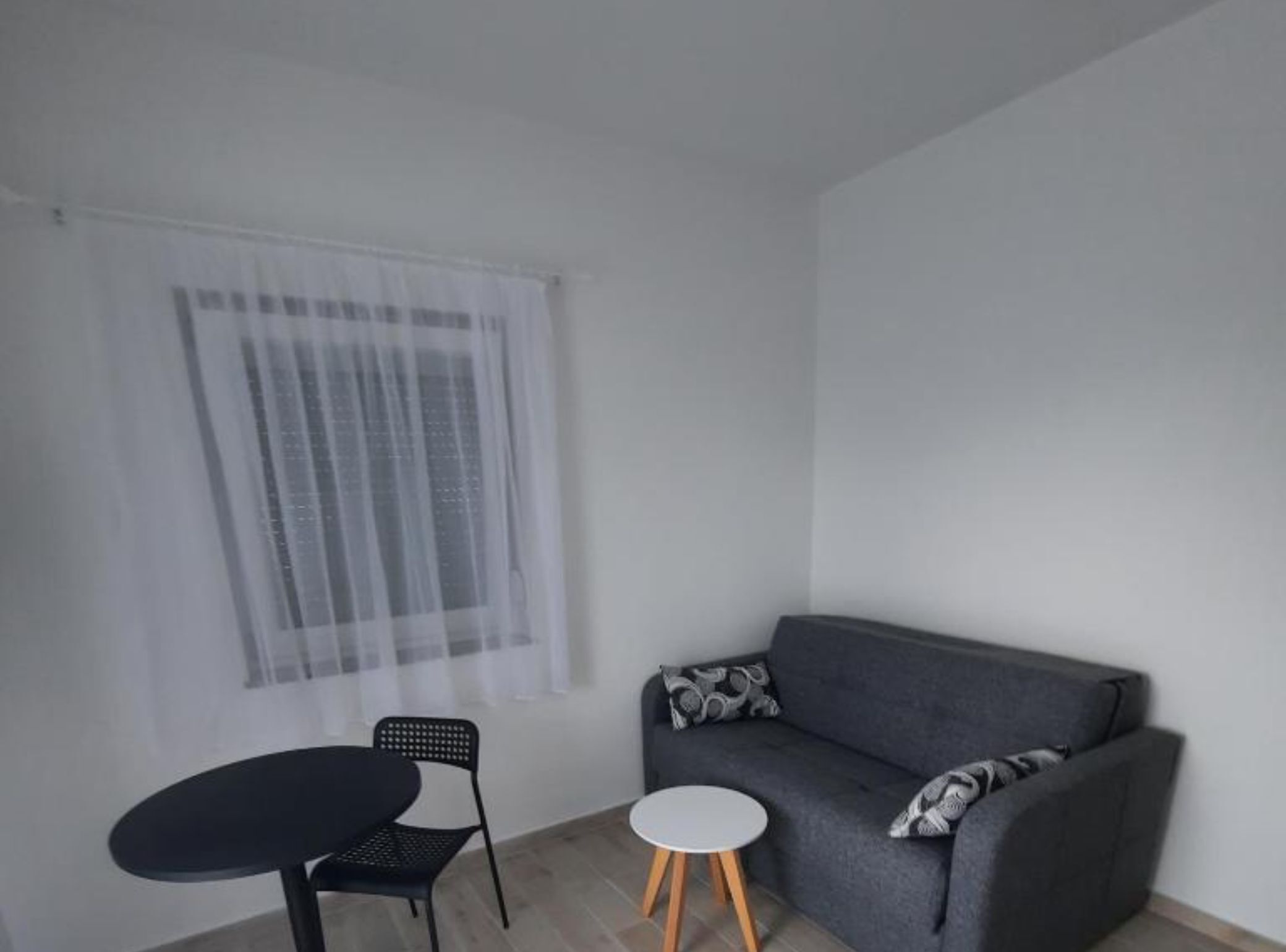 A small, sparsely furnished room with a grey sofa, a black chair, and a small round table.