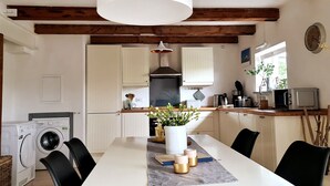 A kitchen with wooden beams, a washing machine, a dining table with chairs, a hanging light fixture, and a clock on the wall. - Apartment 'Nordsee- Ferienwohnung Im Speicher' with Wi-Fi (Meldorf)