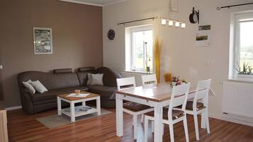 Ein modernes Wohnzimmer mit einer Couch, einem Couchtisch, einem Esstisch mit Stühlen, einem Fenster mit Vorhängen und einer Wanduhr.
