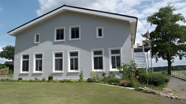 Una casa de dos pisos con techo plano, fachada blanca, numerosas ventanas y un pequeño jardín.