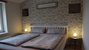 Un dormitorio con pared de ladrillo, cama con dos almohadas, lámparas de noche y un cuadro enmarcado en la pared.