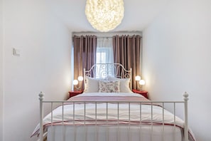 En pænt redt seng med hvid metalramme, to sengeborde med lamper, en dekorativ loftlampe og et vindue med gardiner.