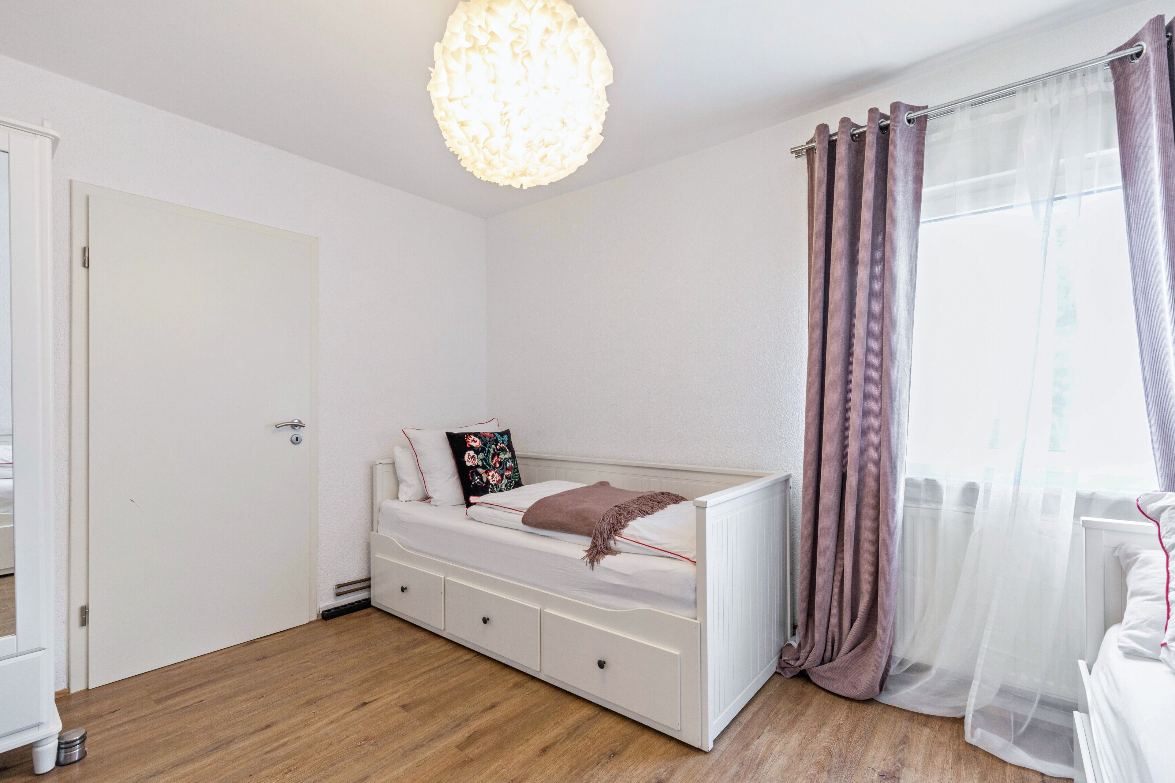 Un dormitorio con cama blanca, mesita de noche, lámpara colgante, ventana con cortinas y puerta.