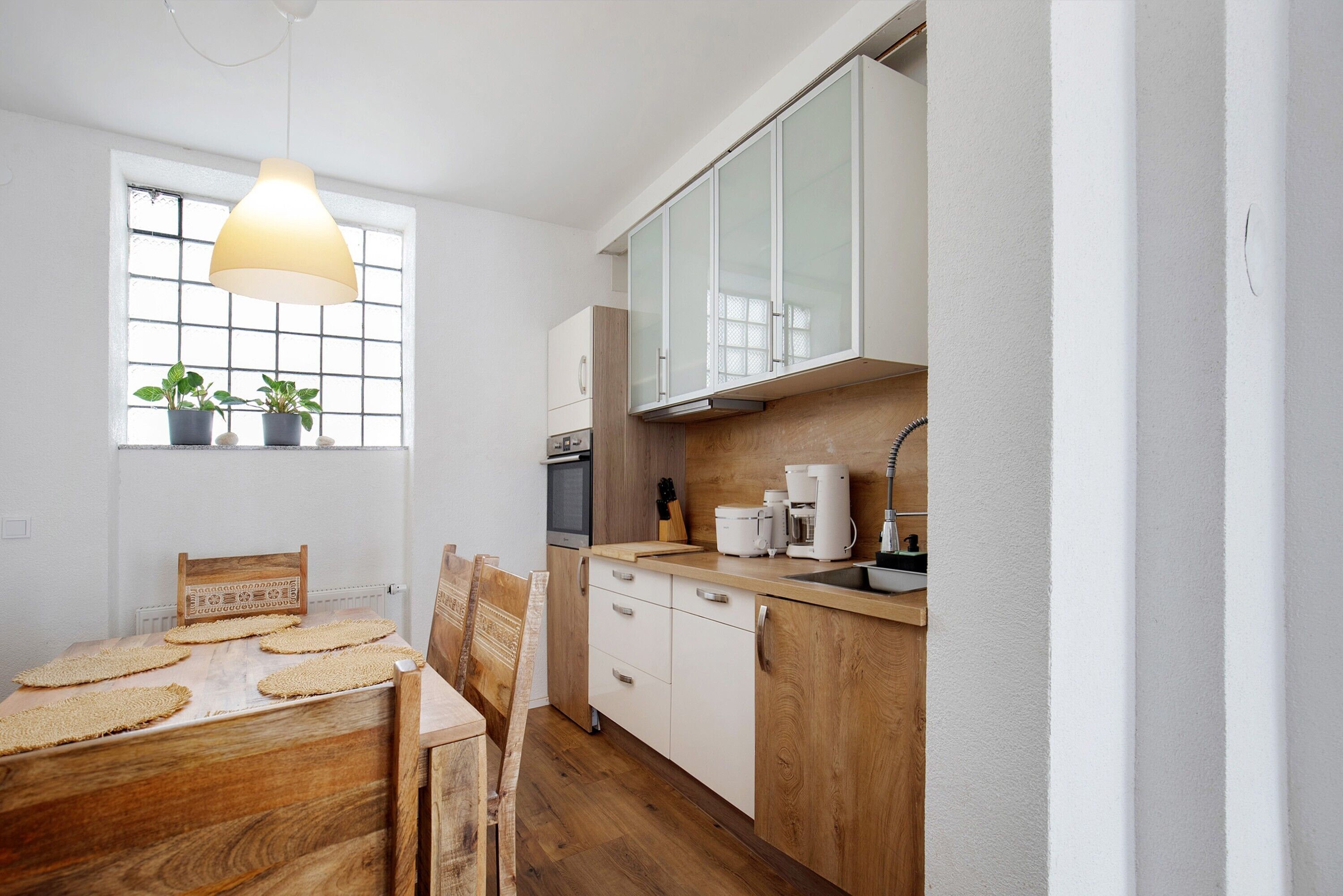 A kitchen with a wooden table, white cabinets, a window with plants, and a hanging light fixture.