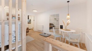 Un interior moderno con un panel de madera, muebles blancos, una mesa redonda con sillas, una lámpara colgante y un radiador.