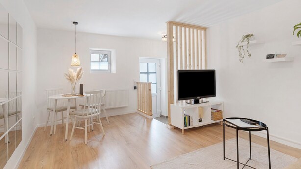 Una sala de estar moderna con piso de madera, paredes blancas, un televisor, una mesa de comedor con sillas y una mesita auxiliar.