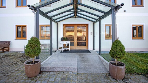 A building entrance with a glass canopy, a wooden door, and two potted trees. - Apartment 10 – Two-Level Living with Separate Bedroom and Well-Equipped Kitchen (Kirchseeon)