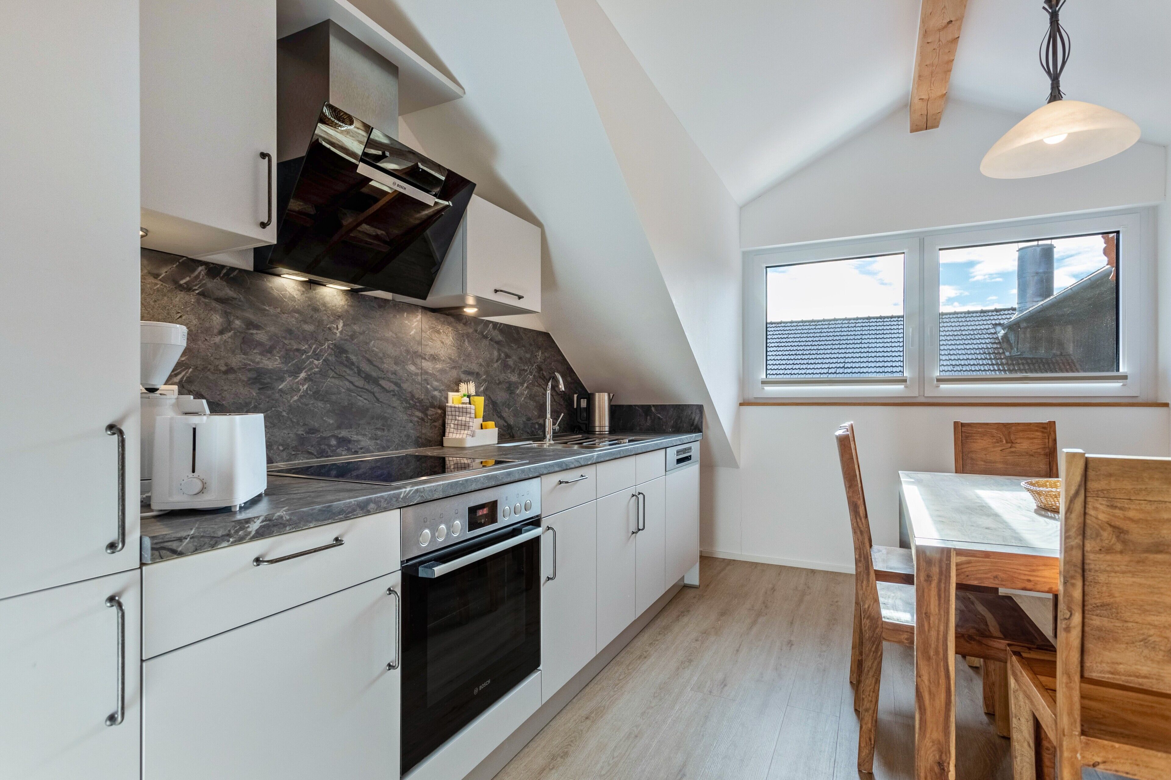 Une cuisine moderne avec des meubles blancs, un four noir, un plan de travail en marbre et un coin repas avec des chaises en bois.