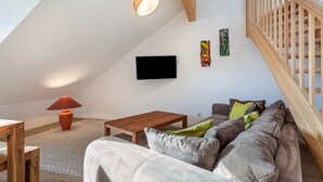 An attic room with a wooden staircase, a flat-screen TV, a sofa with cushions, a wooden coffee table, and a floor lamp. - Apartment 10 – Two-Level Living with Separate Bedroom and Well-Equipped Kitchen (Kirchseeon)