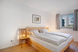 A bedroom with a wooden bed, a nightstand with a lamp, a framed picture on the wall, and a window with curtains.