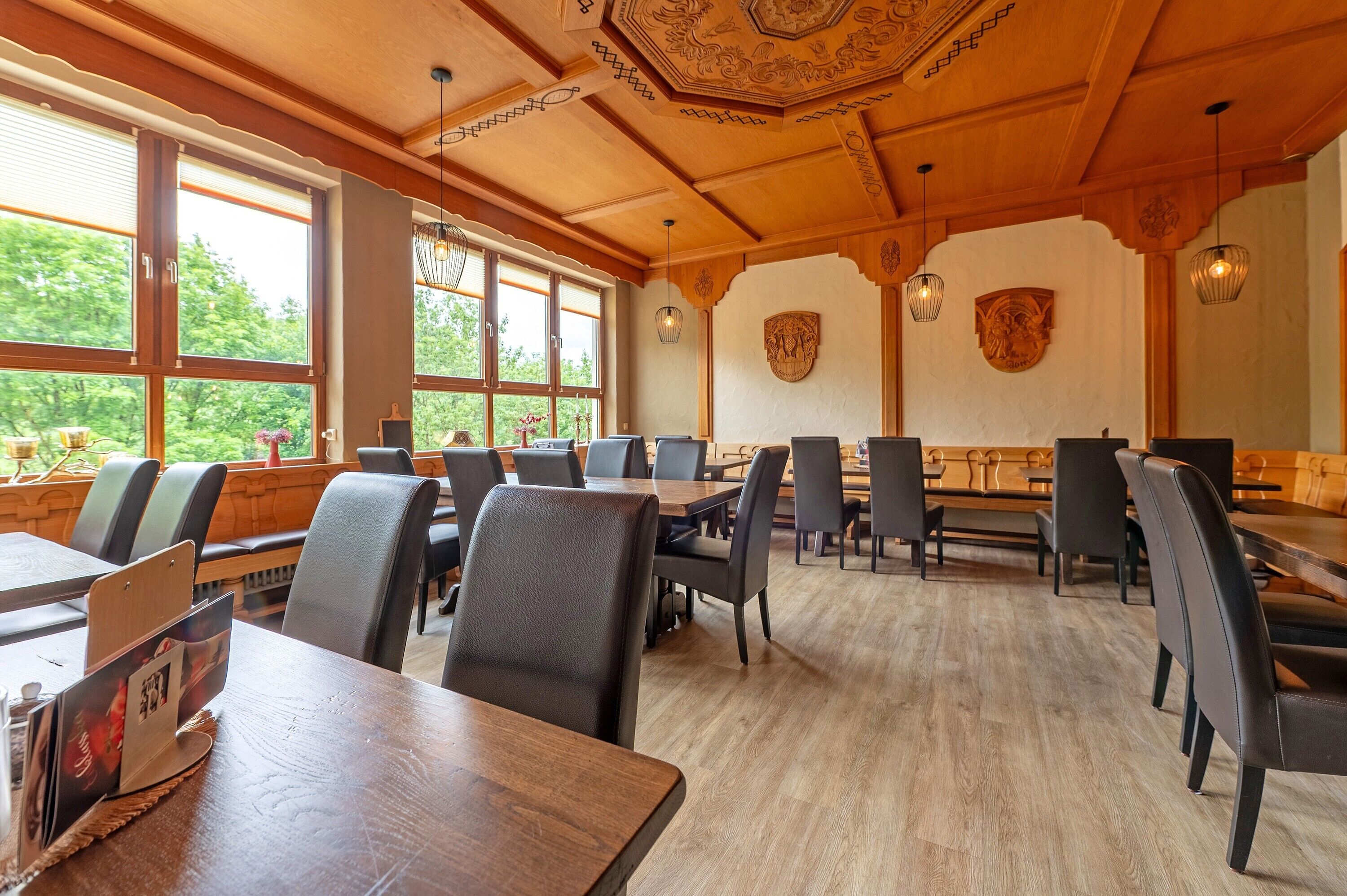 A restaurant with wooden tables and chairs, large windows with views of greenery, and decorative wall art.