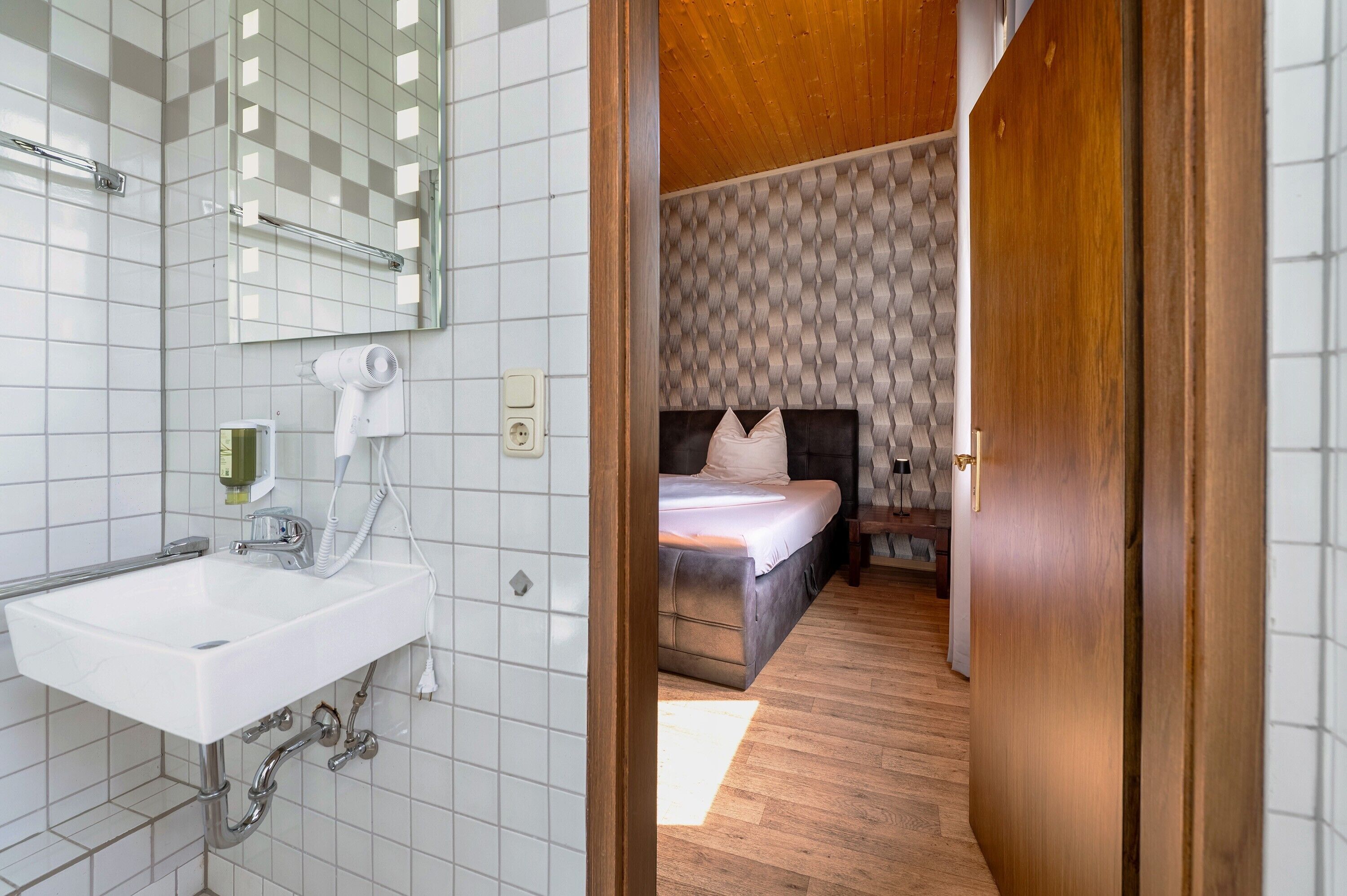 A bathroom with a white sink, mirror, and hairdryer, and a bedroom with a bed, wooden floor, and patterned wallpaper.