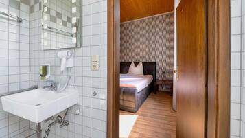 A bathroom with a white sink, mirror, and hairdryer, and a bedroom with a bed, wooden floor, and patterned wallpaper.