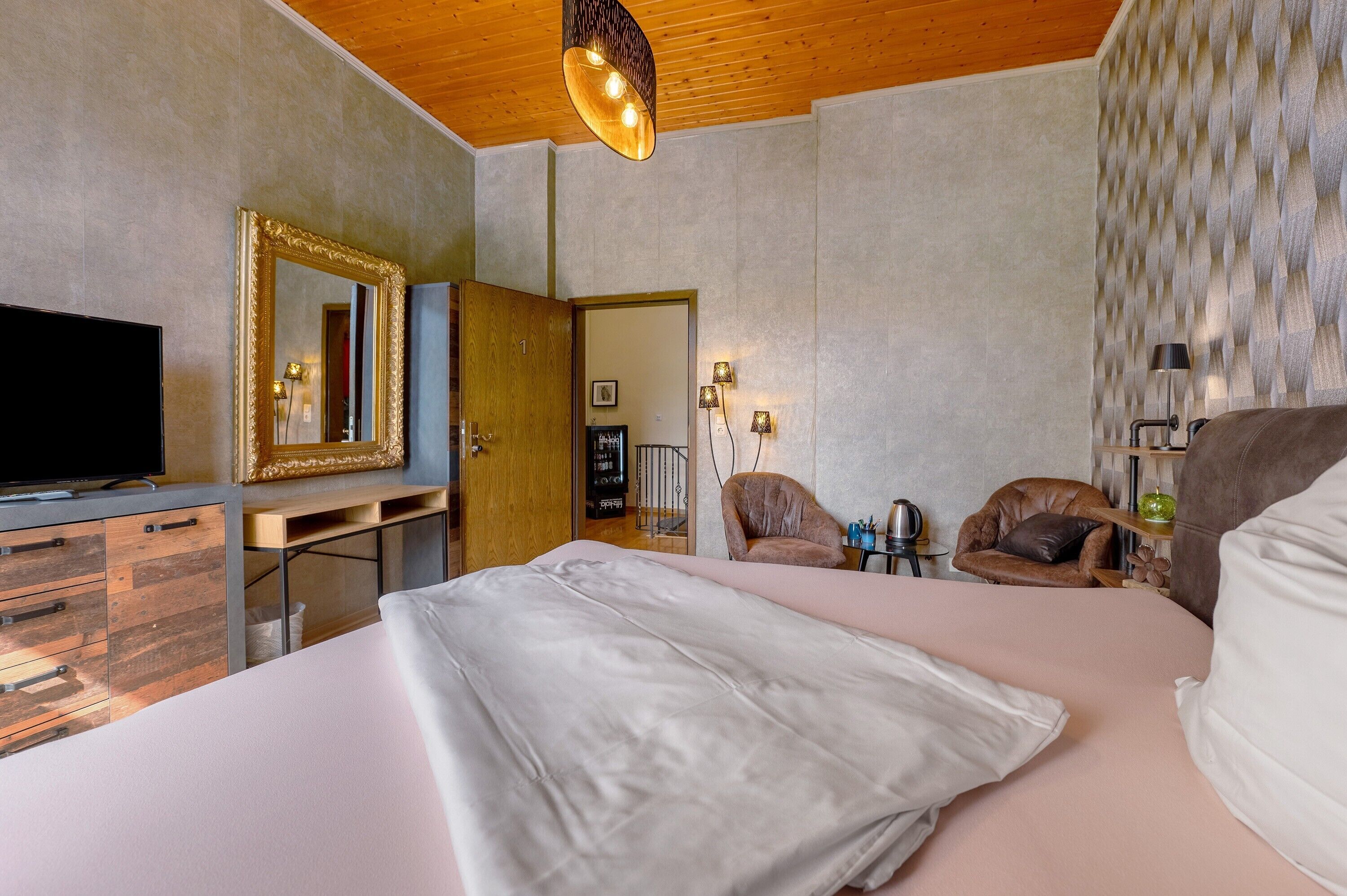 A bedroom with a bed, a TV, a dresser, a mirror, and a door leading to another room.