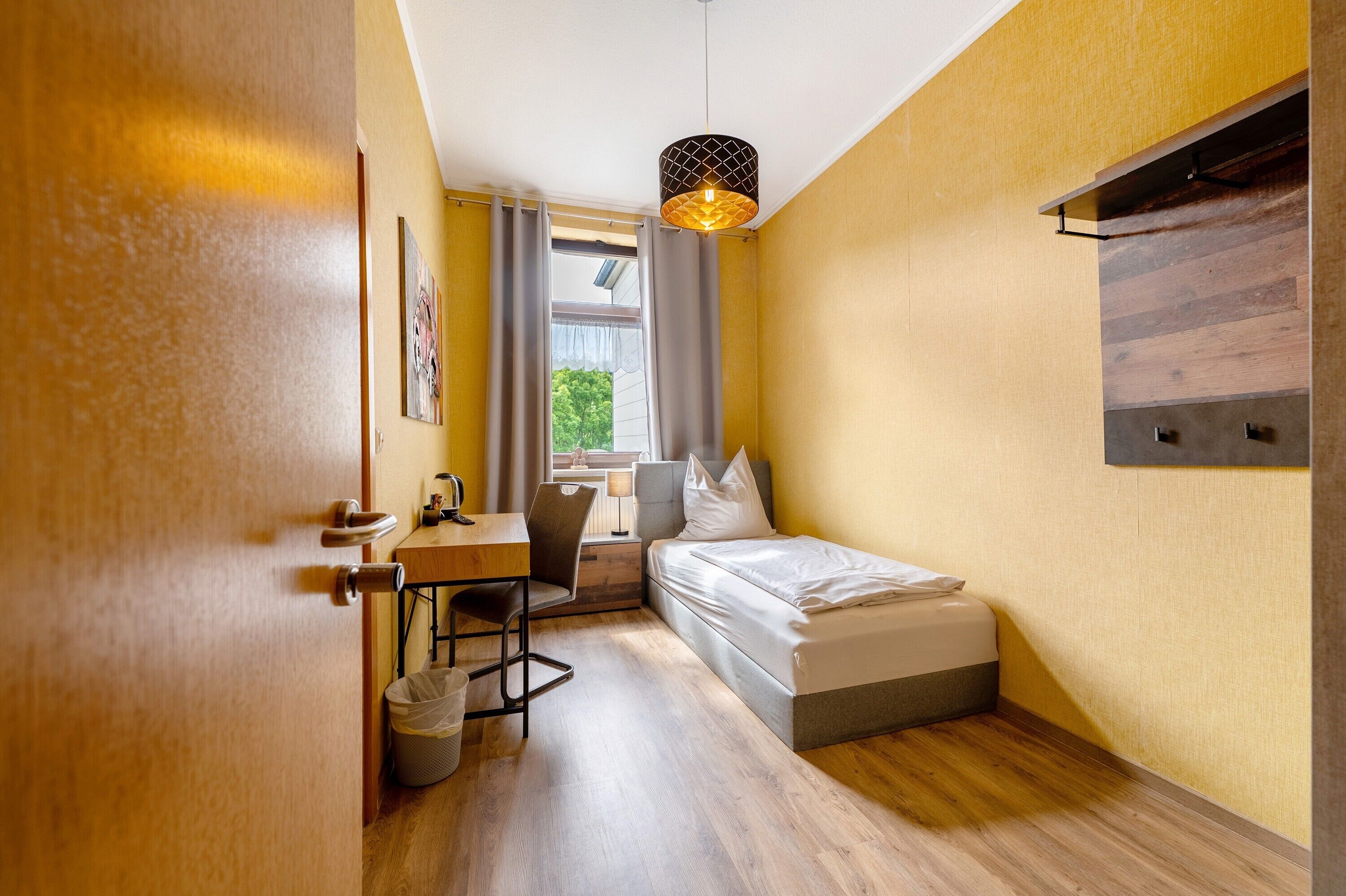 Ett litet hotellrum med en säng, ett skrivbord, en stol och en hängande lampa.