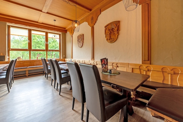 Un espace repas avec des tables et des chaises en bois, de grandes fenêtres équipées de stores et des œuvres d’art décoratives sur les murs.