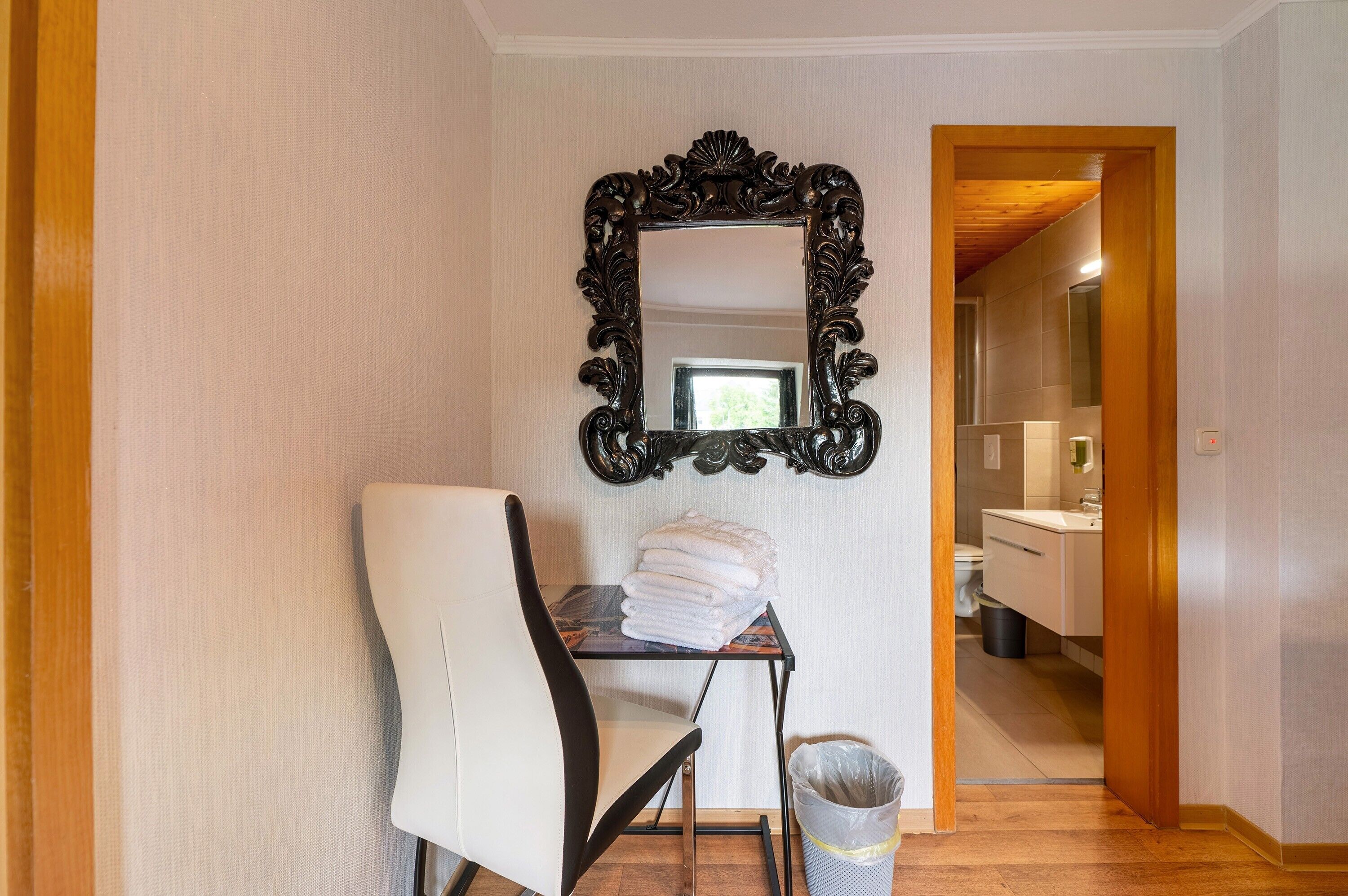 Ein Hotelzimmer mit einem großen, verzierten Spiegel, einem weißen Sessel, einem kleinen Tisch mit gefalteten Handtüchern und einem sichtbaren Badezimmer im Hintergrund.