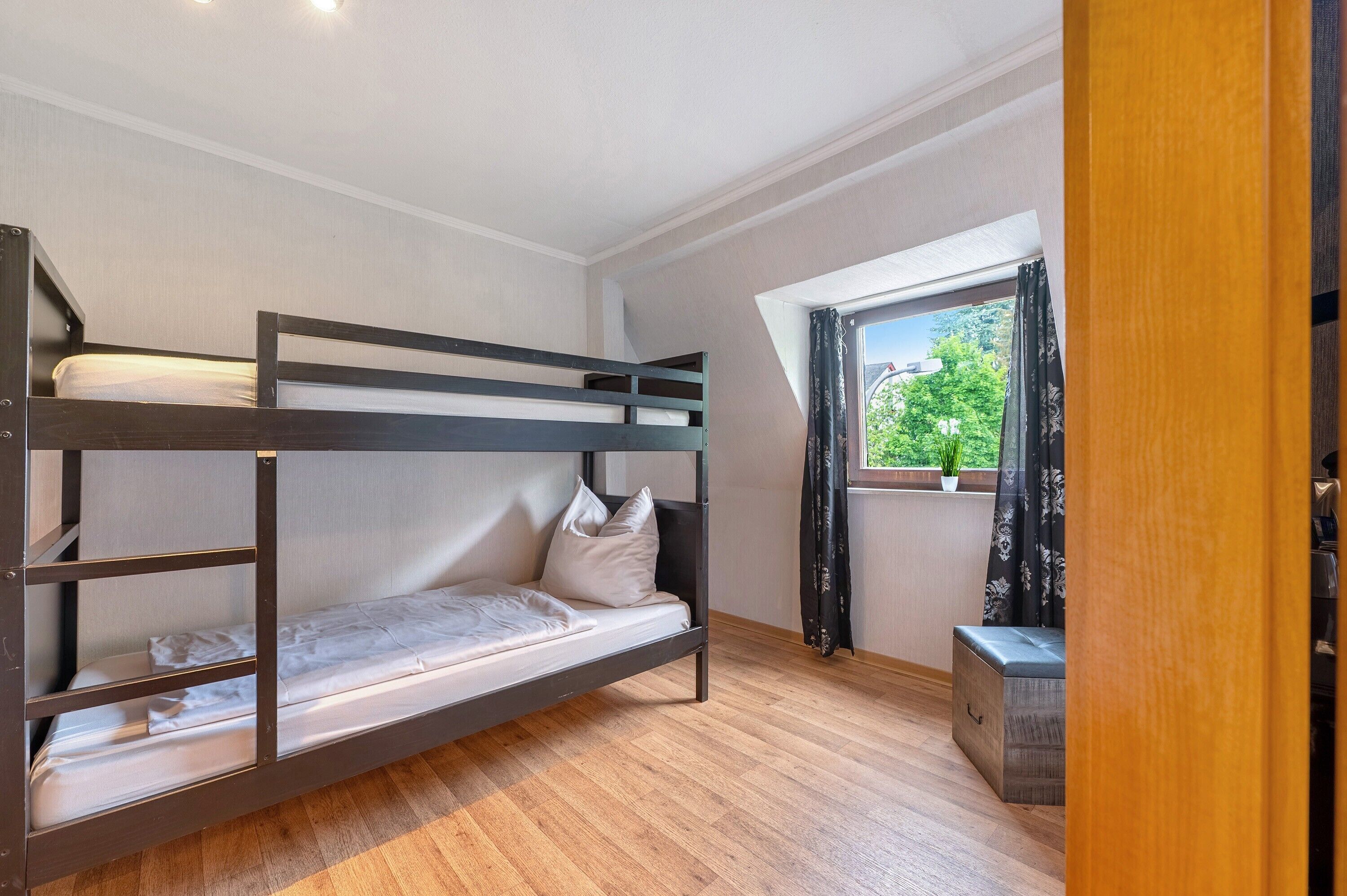 A bunk bed room with a wooden floor, a window with curtains, and a small plant on the windowsill.