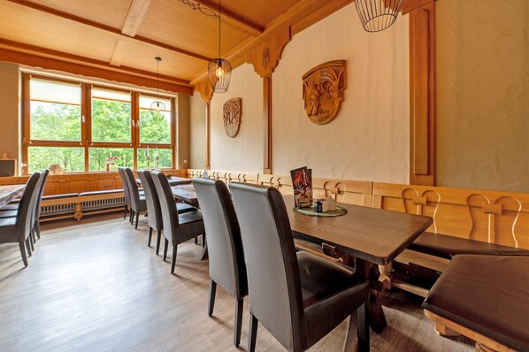 Un espace repas avec des tables et des chaises en bois, de grandes fenêtres équipées de stores et des œuvres d’art décoratives sur les murs.