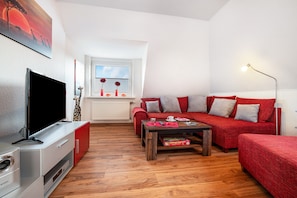 A modern living room with a flat-screen TV, red sofas, a wooden coffee table, and a window with a view.