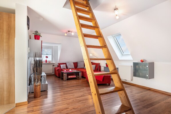 A loft-style room with a wooden ladder, a red sofa, a dining table, and a window.