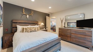 A bedroom with a large bed, wooden headboard, a mounted deer skull, a dresser, a television, and a mirror.