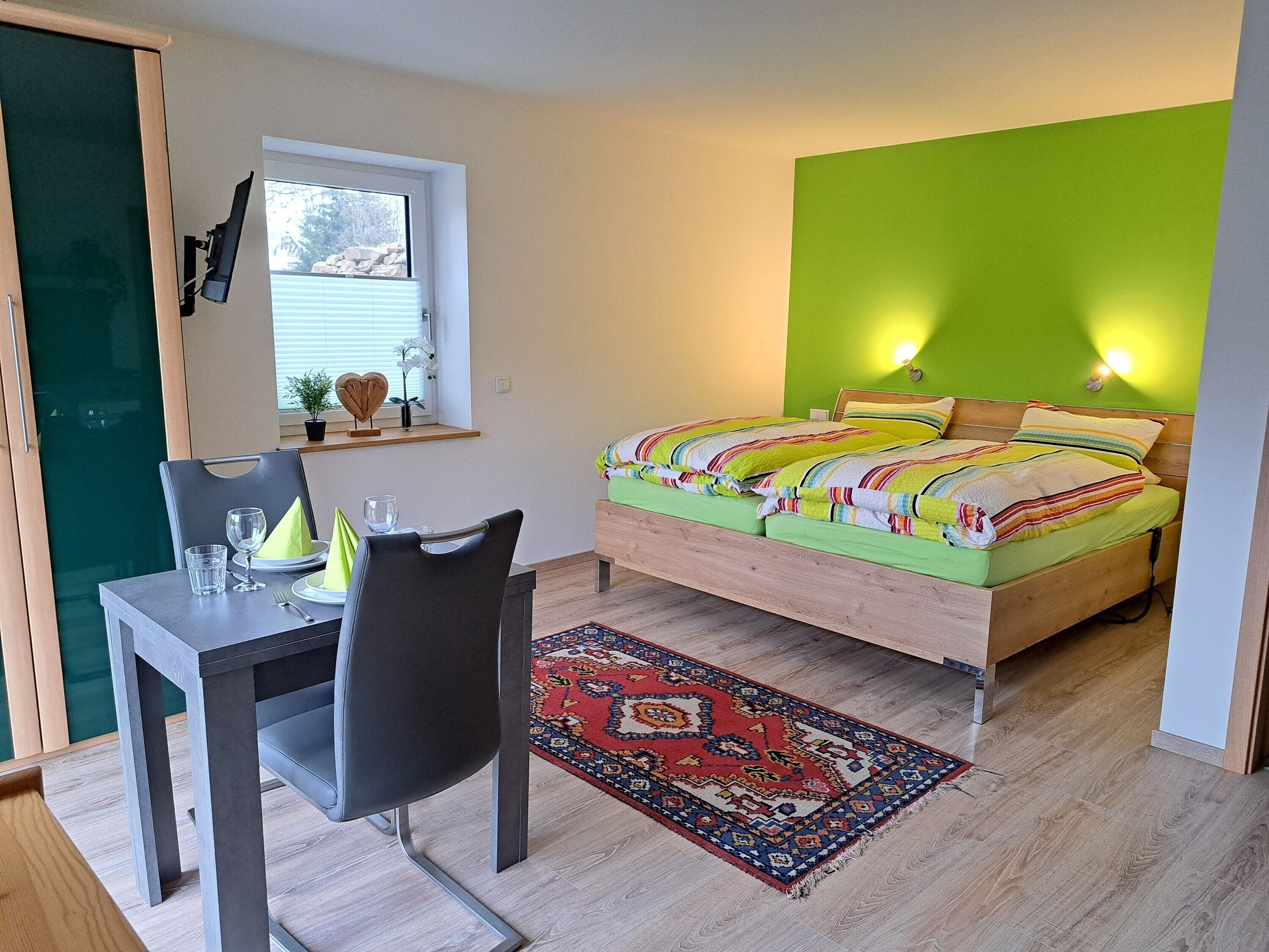 A room with a bed, a dining table with chairs, a window, and a green wall.