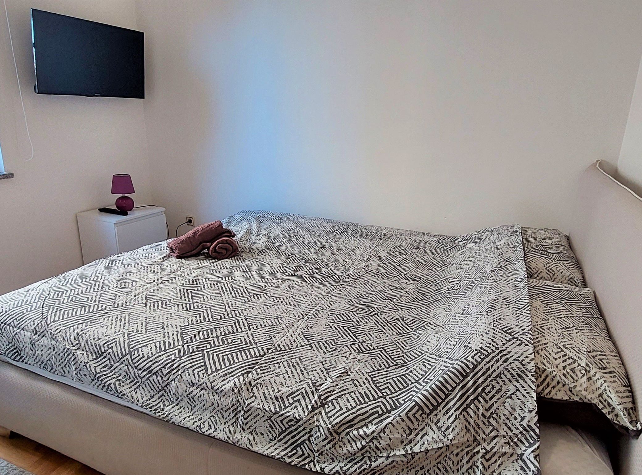 A neatly made bed with a patterned bedspread, a folded towel, a bedside lamp, and a flat-screen TV mounted on the wall.