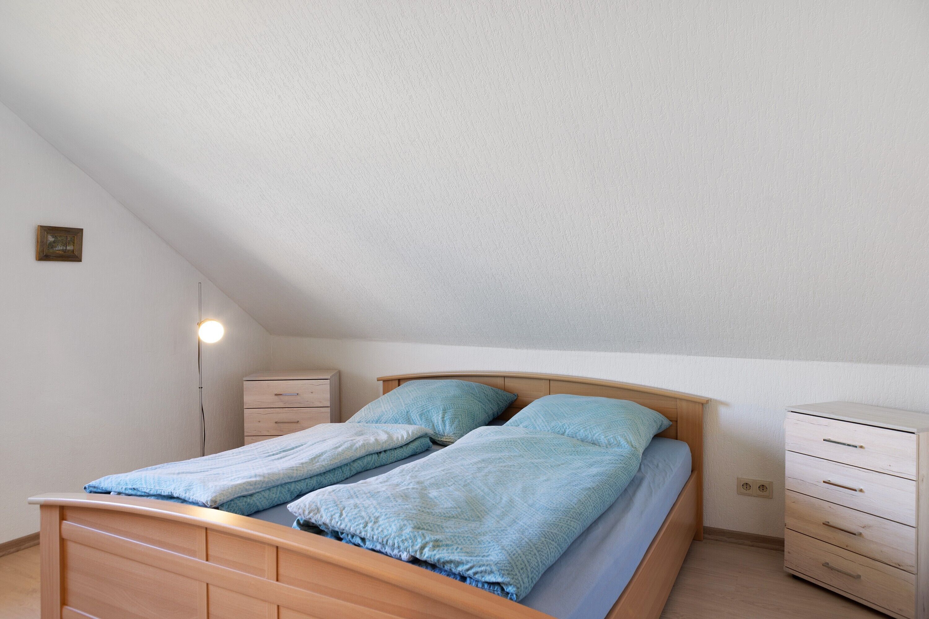 A single bed with a wooden frame, a bedside table, a nightstand, and a dresser in a room with sloped ceilings.