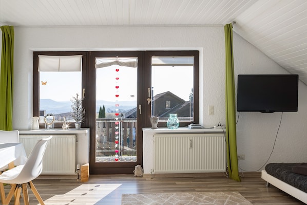 A room with a dining table, a sofa, a TV mounted on the wall, and a view through the glass doors.