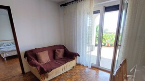 A room with a wooden parquet floor, a maroon sofa with cushions, a bed with a white bedspread, and a sliding glass door leading to an outdoor area. - Sea View Apartment Crikvenica With Terrace (Crikvenica)