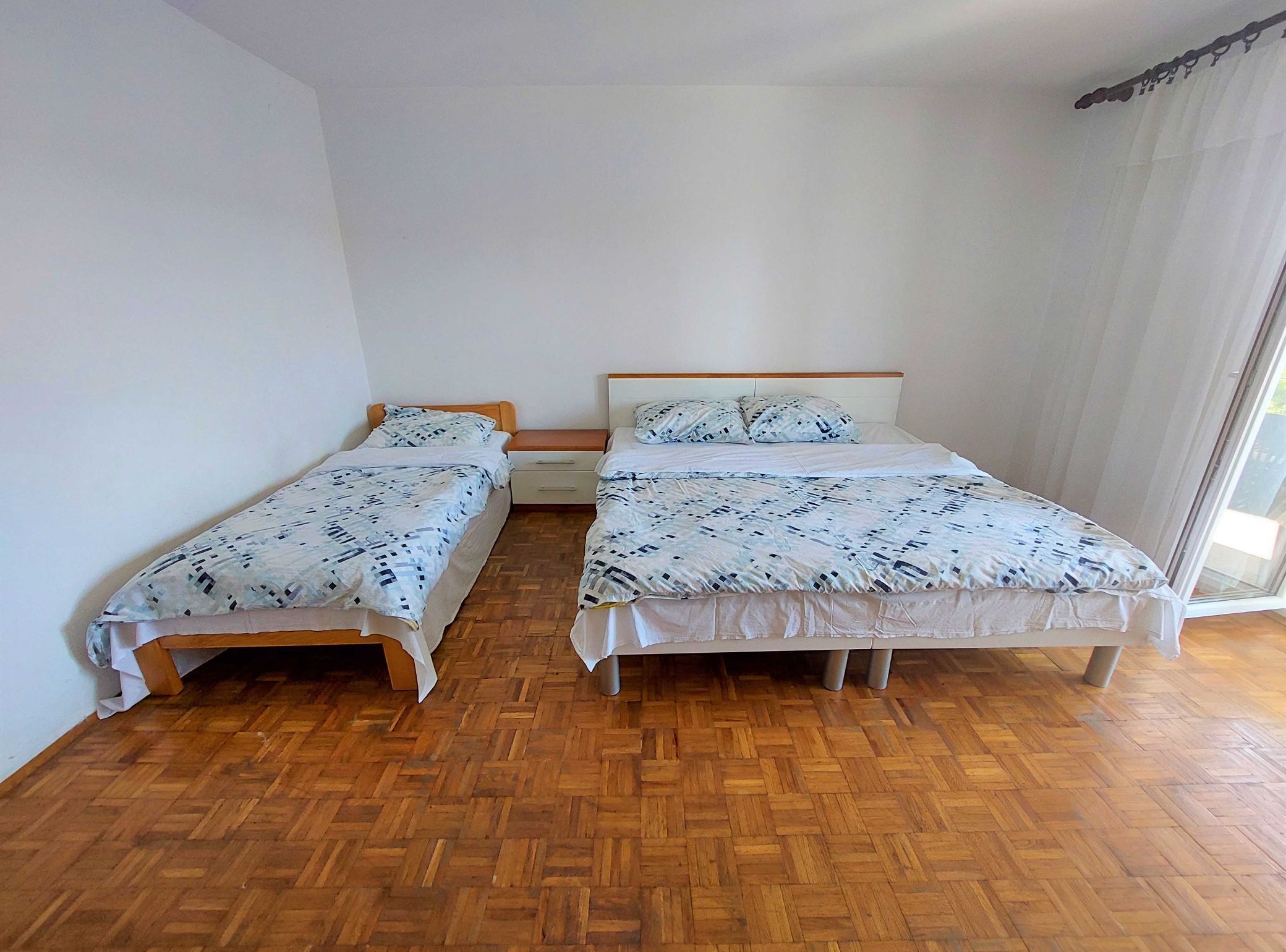 A room with two single beds, a wooden floor, and a large window with curtains.