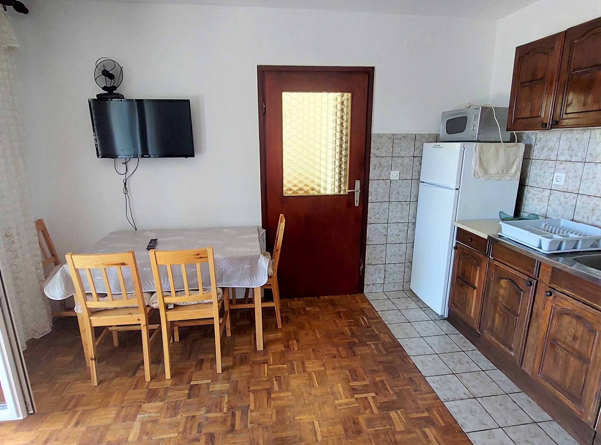 A compact kitchen with a wooden table and chairs, a microwave, a refrigerator, and a TV mounted on the wall.