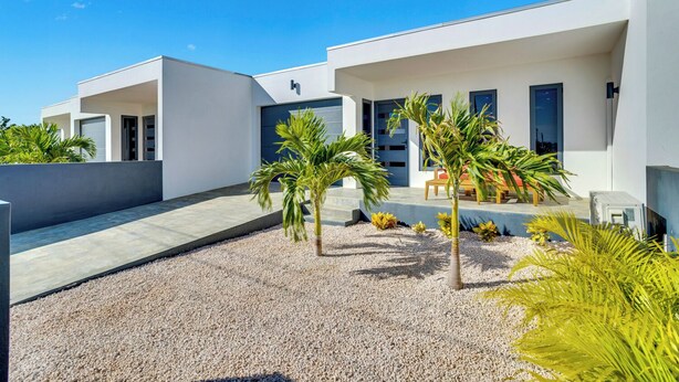 Una casa moderna de dos pisos con un patio que incluye palmeras y un espacio para sentarse.