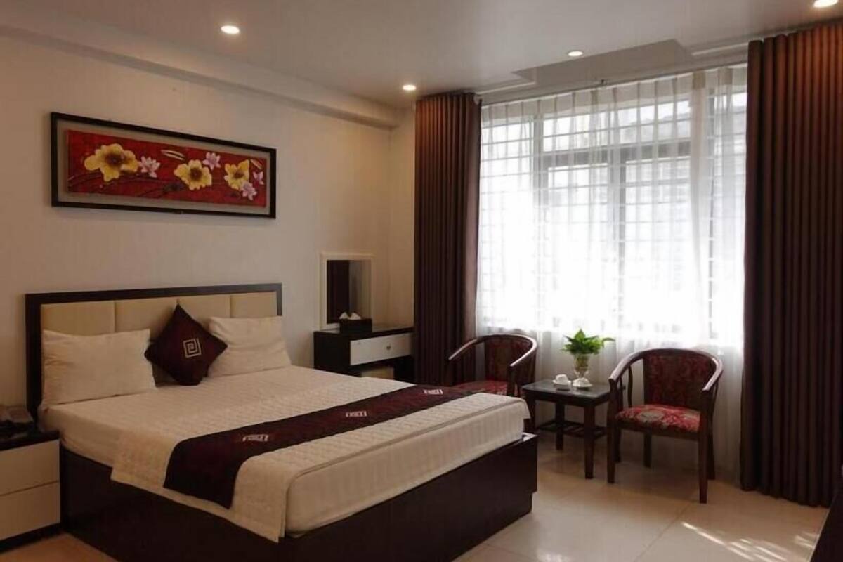 A hotel room with a large bed, a desk, a chair, a window with curtains, and a painting on the wall.