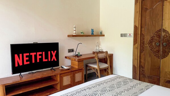 A hotel room with a wooden desk, a chair, a television displaying the Netflix logo, a bedside table, and a patterned bedspread. - Leafy Home Canggu (Canggu)
