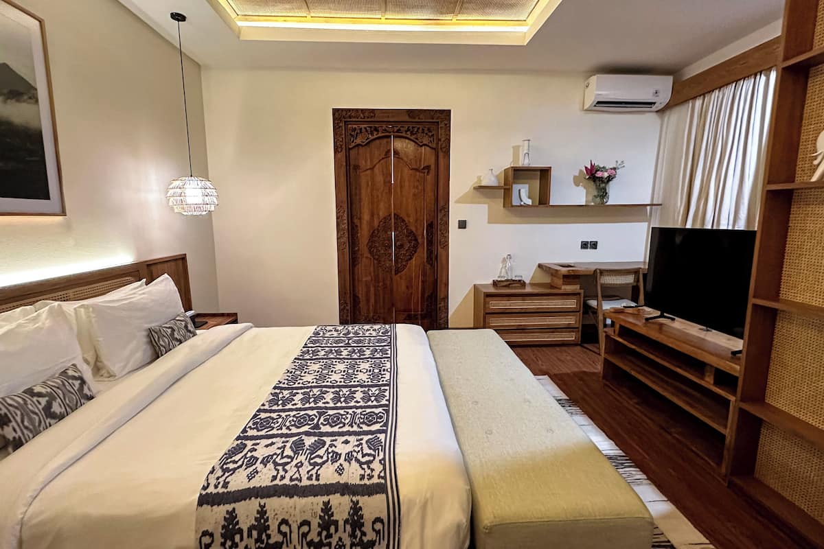 A well-lit bedroom with a large bed, a wooden wardrobe, a television, and a shelf with decorative items.