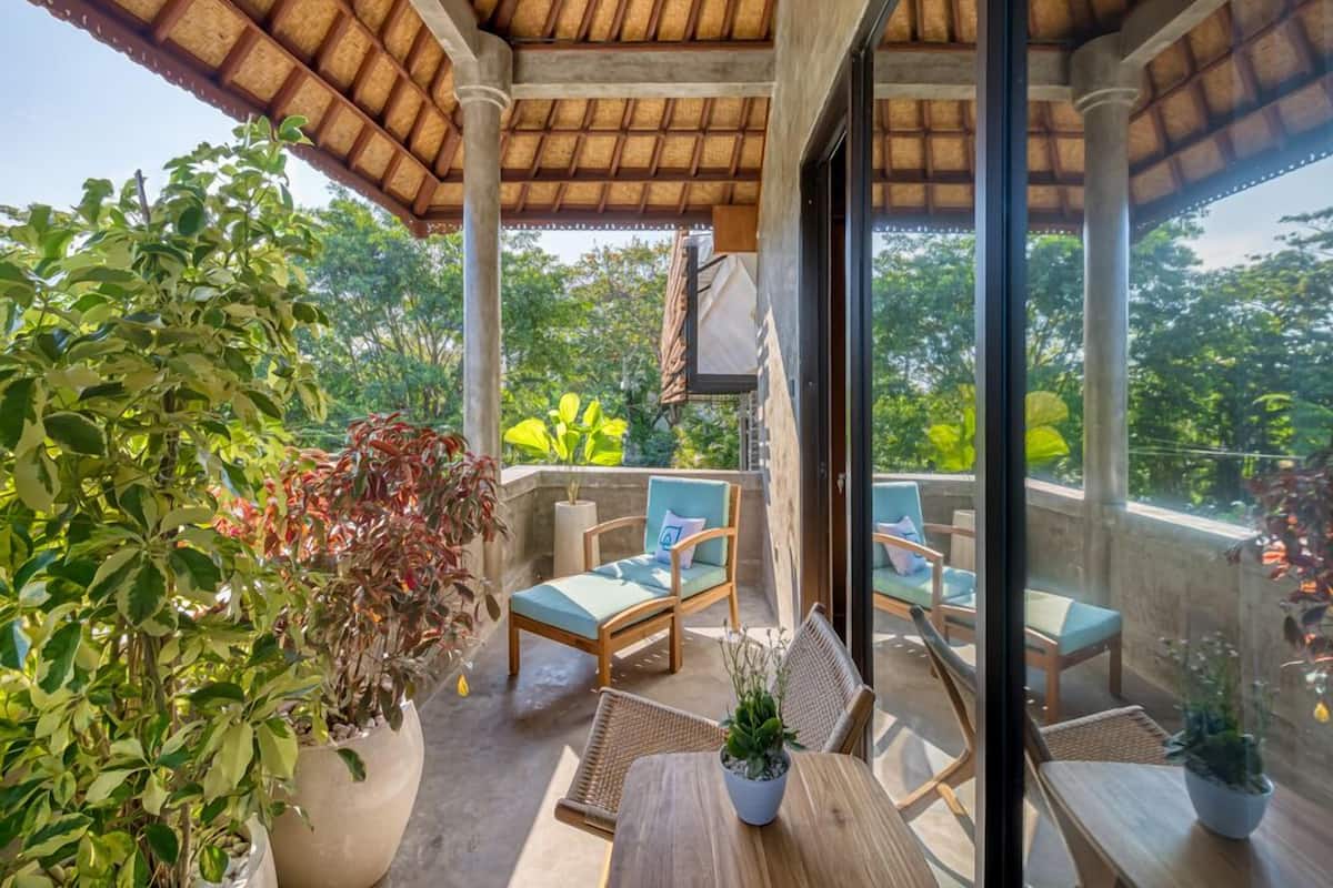 A covered balcony with wooden chairs and tables, surrounded by potted plants and a view of greenery.