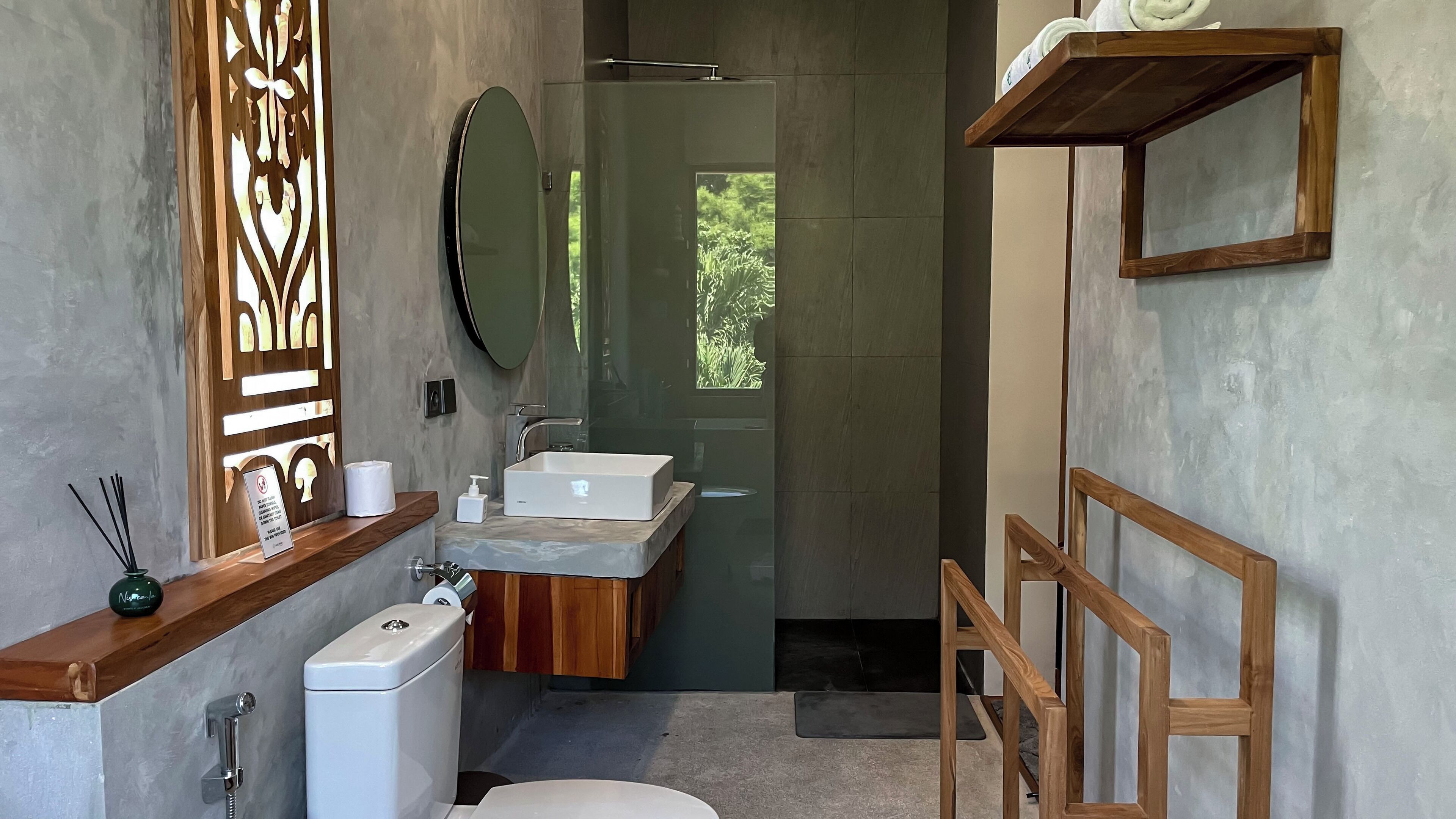 A modern bathroom with a toilet, sink, and shower. There is a wooden shelf with toiletries and a mirror above the sink.